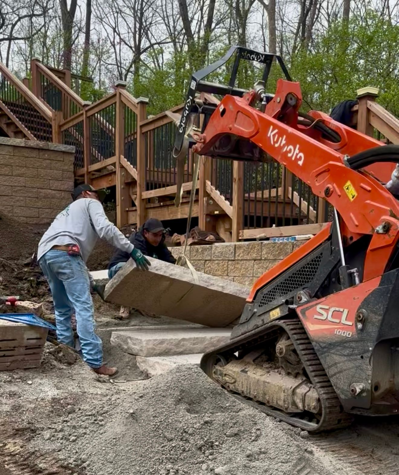 Two workers using a Kubota machine to place a stone step near stairs.