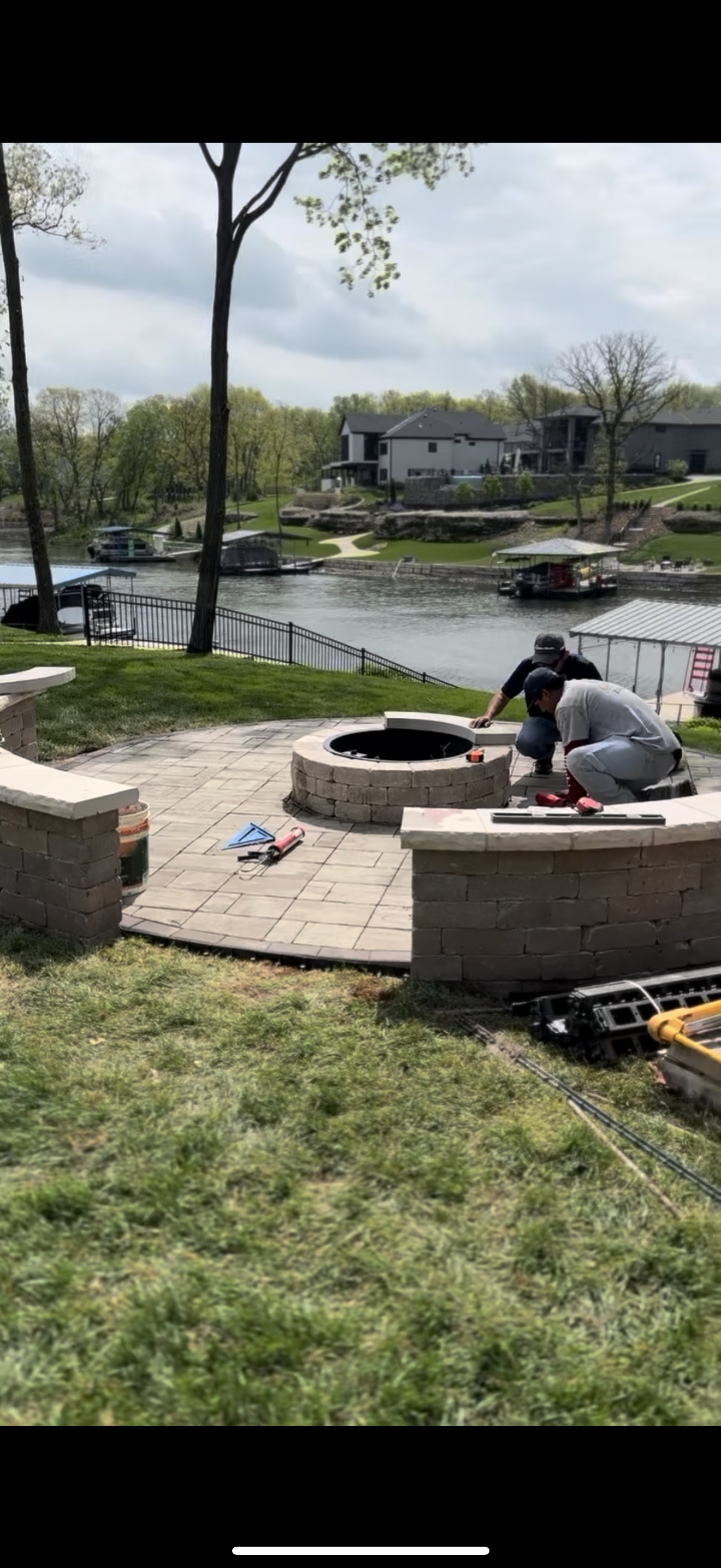 Two people working on a stone fire pit, overlooking a lake with houses, trees, and docks.