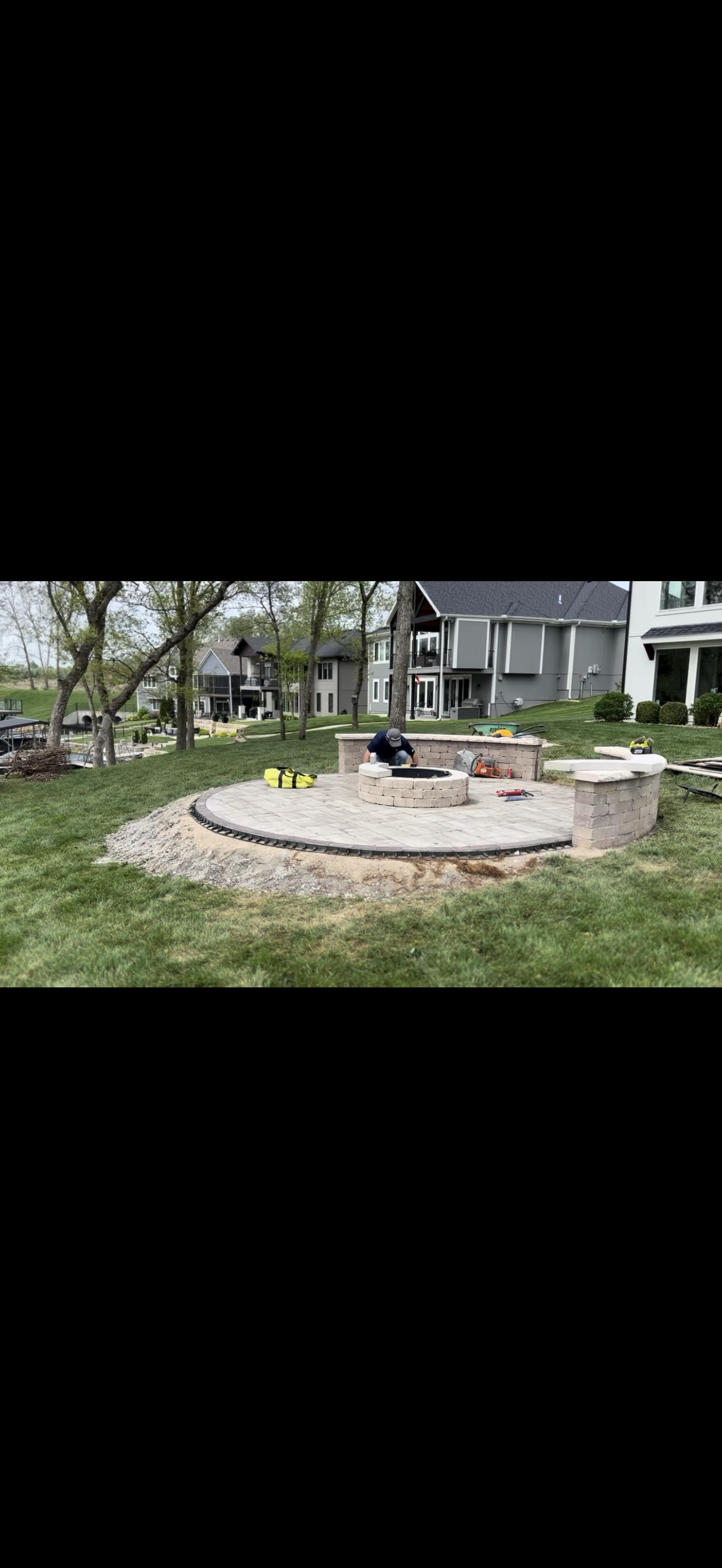 Backyard with a stone fire pit, surrounded by gravel, and seating, in front of houses on a sunny day.