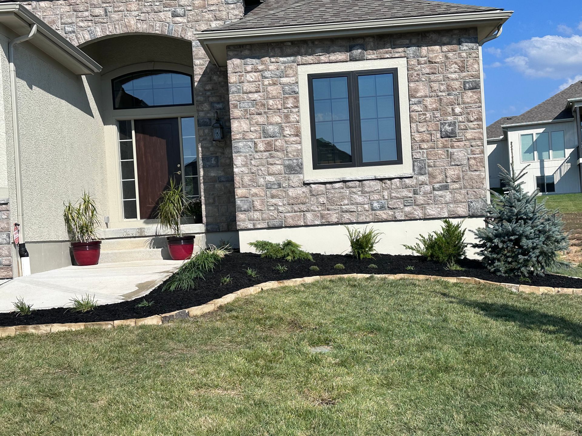 House exterior with stone facade, dark window trim, small shrubs, and mulch bed.
