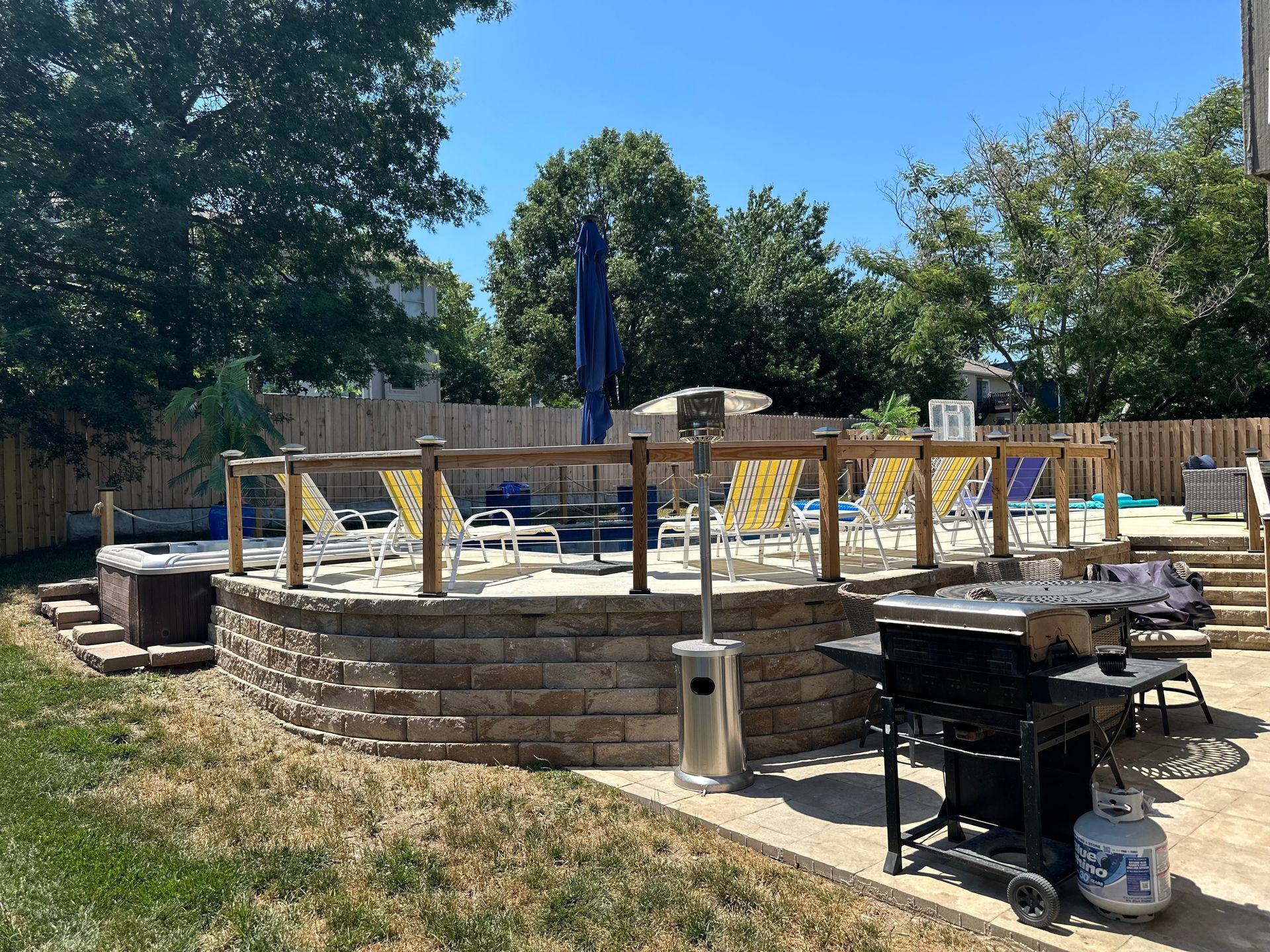 Backyard with a pool, hot tub, grill, and patio furniture under a blue umbrella on a sunny day.