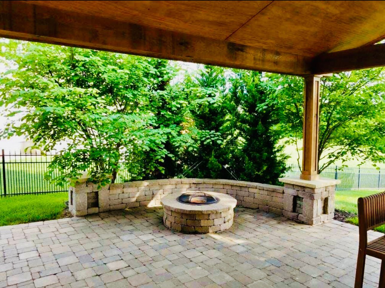 Brick patio with stone fire pit, surrounded by a low stone wall under a wooden pergola, overlooking green trees and a fence.