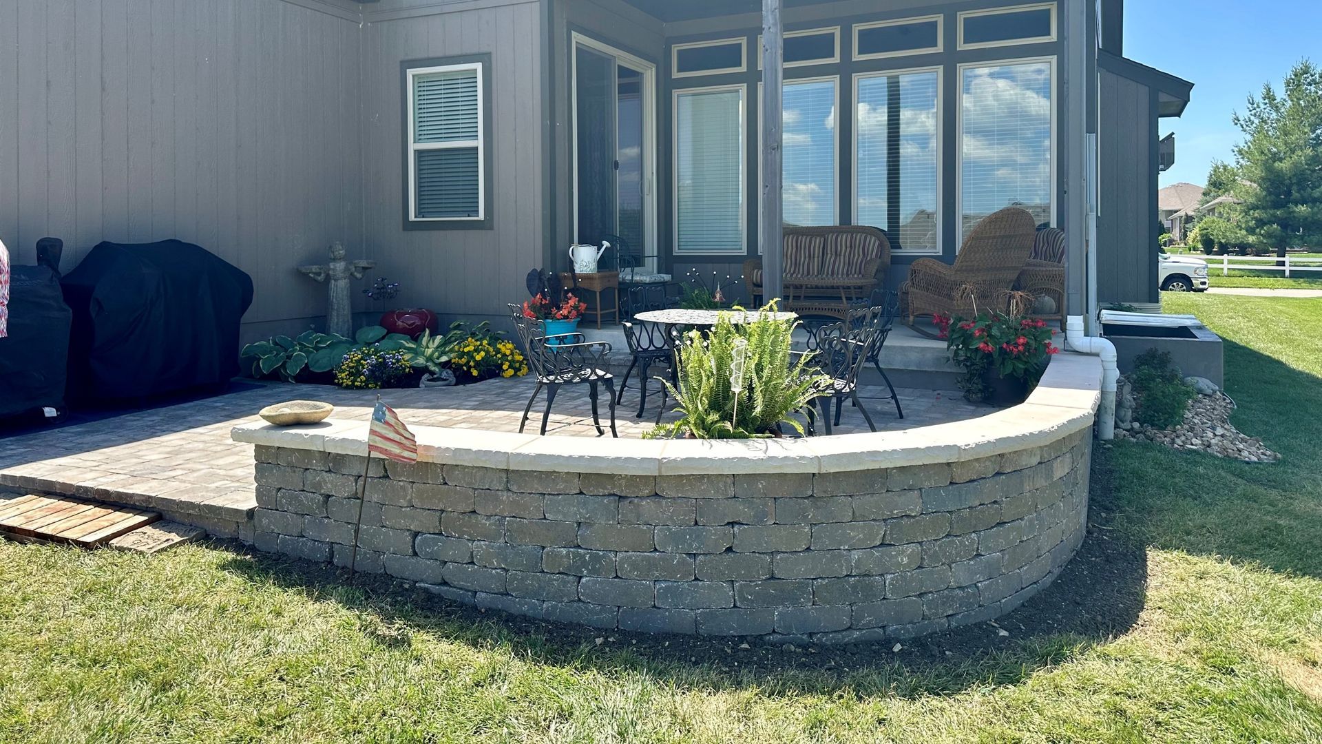Curved brick patio with landscaping, outdoor furniture, and house in the background. Green grass in the foreground.