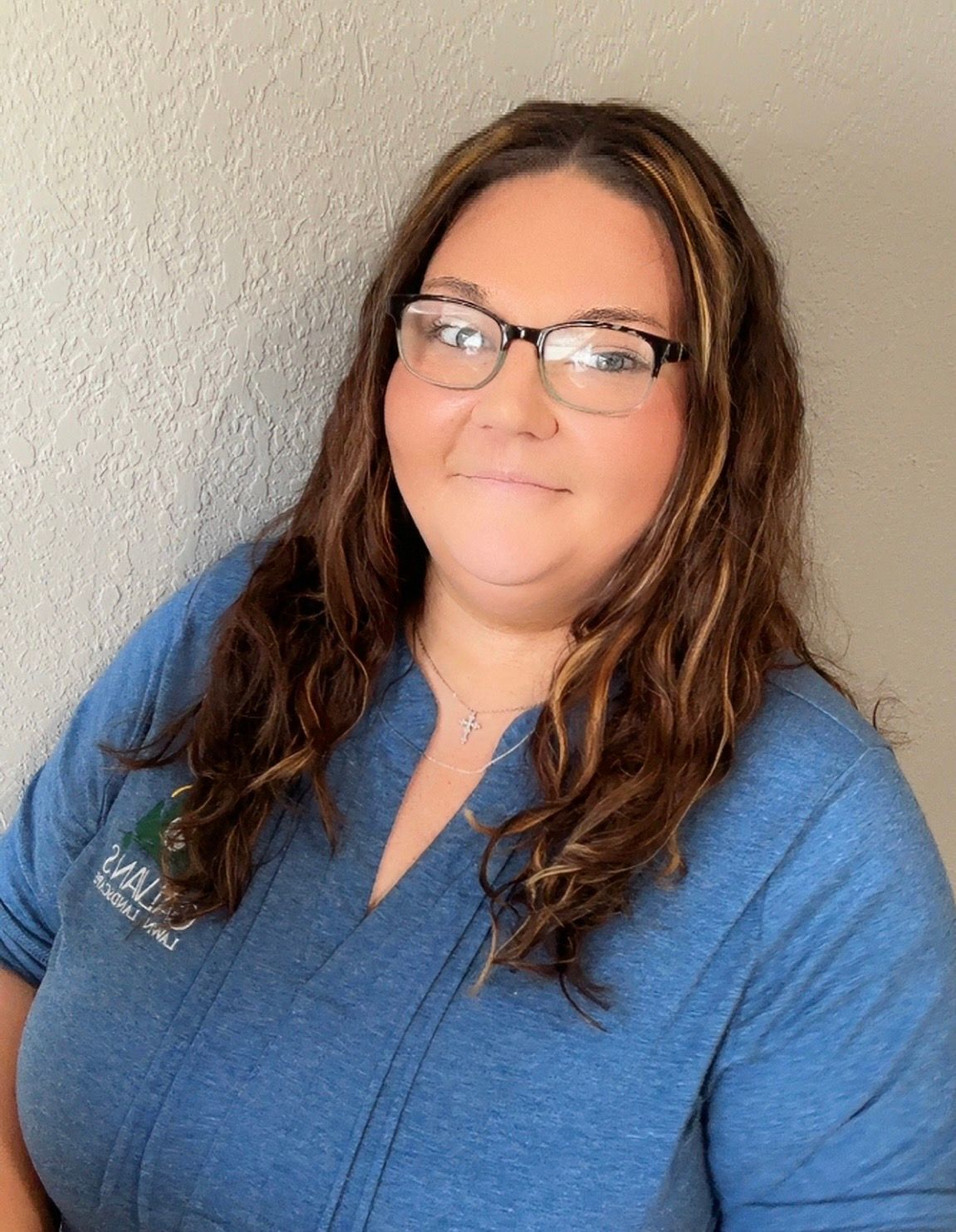 A smiling person with long, wavy brown hair and glasses wearing a blue top against a textured wall.