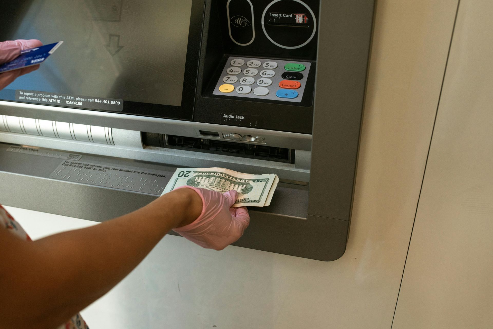 Persona usando un cajero automático, recibiendo efectivo y sosteniendo una tarjeta. Lleva guantes rosas.