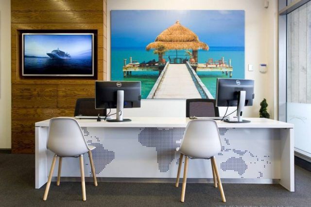 Oficina de agencia de viajes con escritorio, computadoras y obras de arte con escenas de playa.
