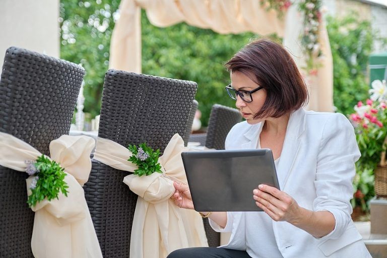 Mujer con blazer blanco revisa una tableta y sillas decoradas en un entorno de boda.