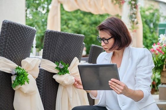 Mujer con blazer blanco revisa una tableta y sillas decoradas en un entorno de boda.