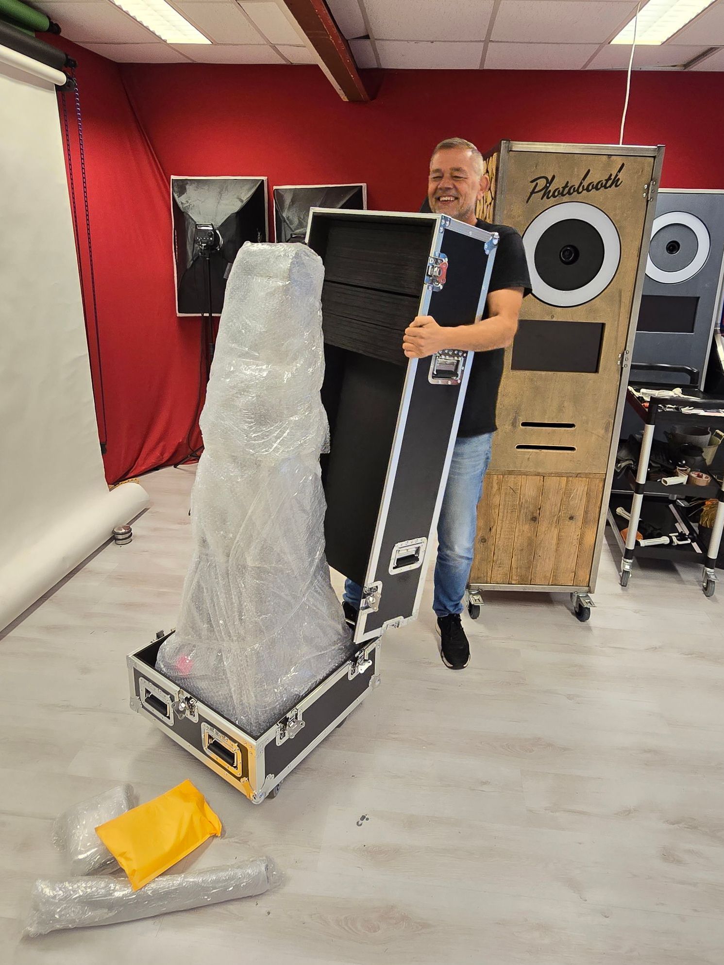Een man pakt apparatuur uit, waarschijnlijk voor een fotohokje, in een roodkleurige ruimte waar meerdere fotohokjes zichtbaar zijn.