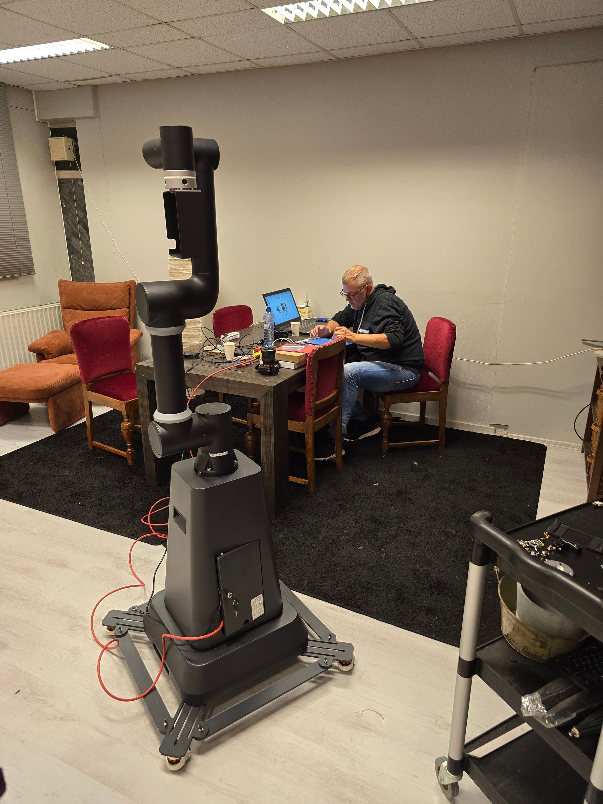 Een hoge robotarm staat naast een man die aan een tafel werkt, in een kamer met een vloerkleed en stoelen.