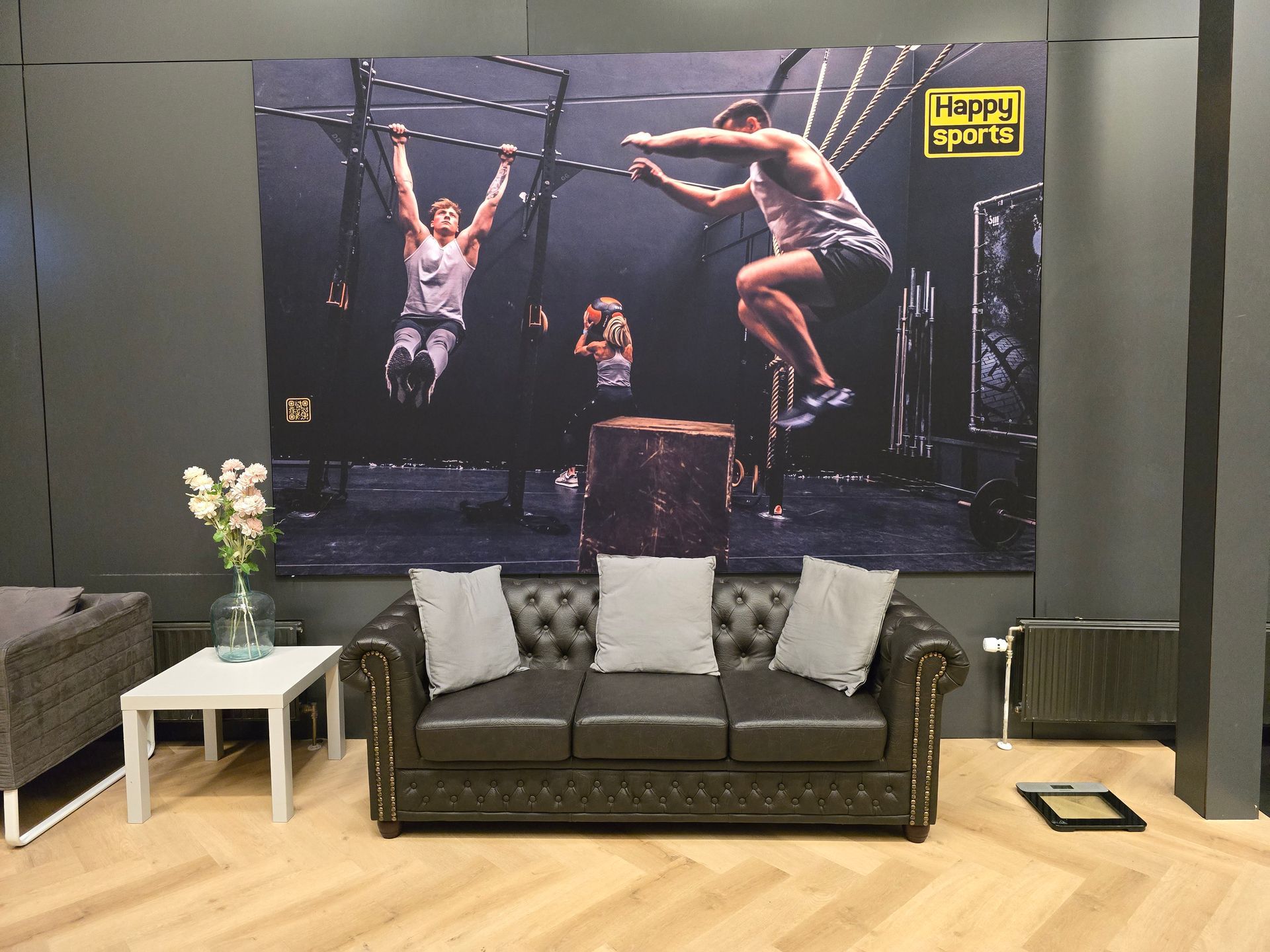 Een bank staat voor een muurschildering met een fitnessthema. Mensen zijn aan het sporten. Ook zijn een witte tafel, een vaas met bloemen en een weegschaal zichtbaar.