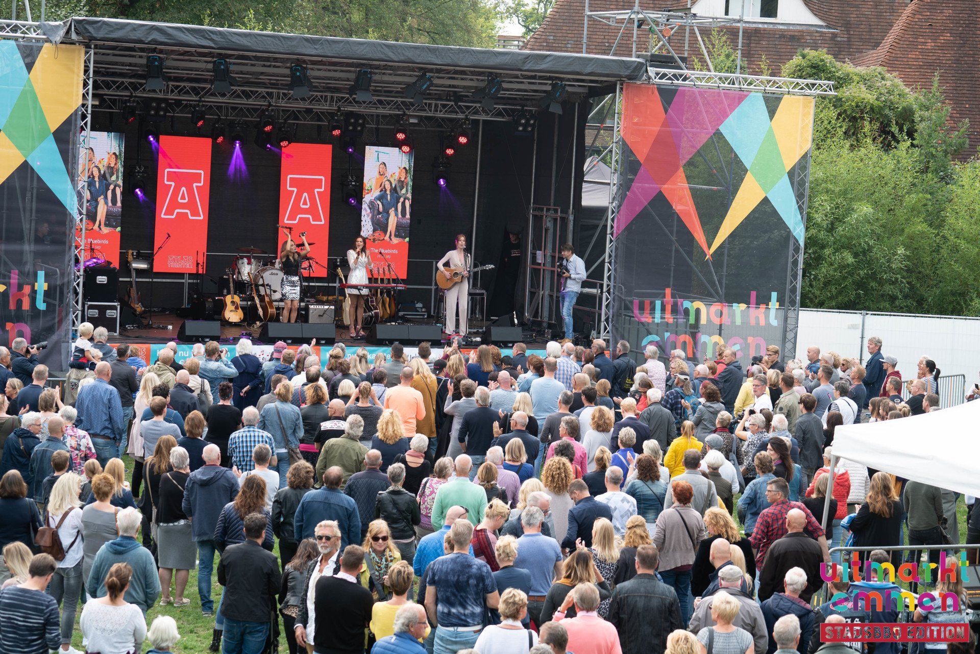 Livemuziek op een podium bij een openluchtevenement met een grote menigte; spandoeken en kleurrijke geometrische versieringen.