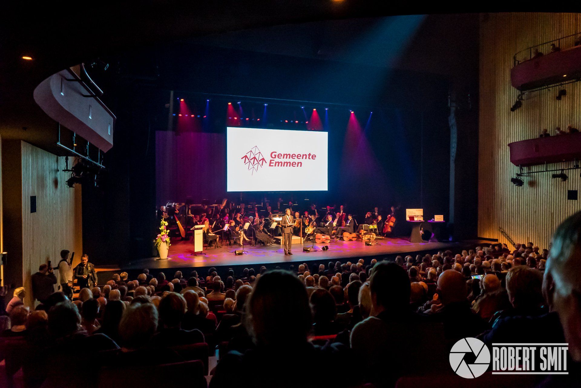 Het publiek kijkt naar een voorstelling op het podium met een orkest en een presentator; op een groot scherm wordt het logo weergegeven.