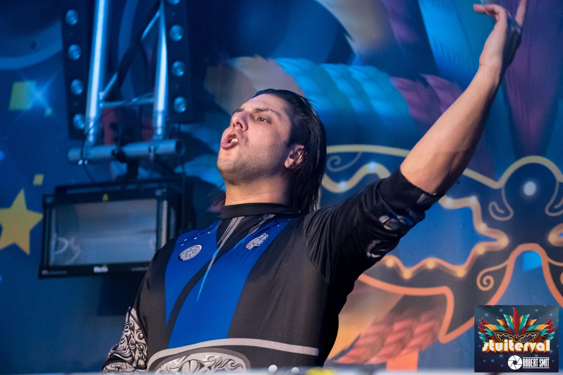 DJ op het podium met opgeheven arm, gekleed in een blauw-zwart pak. Kleurrijk decor, podiumverlichting.