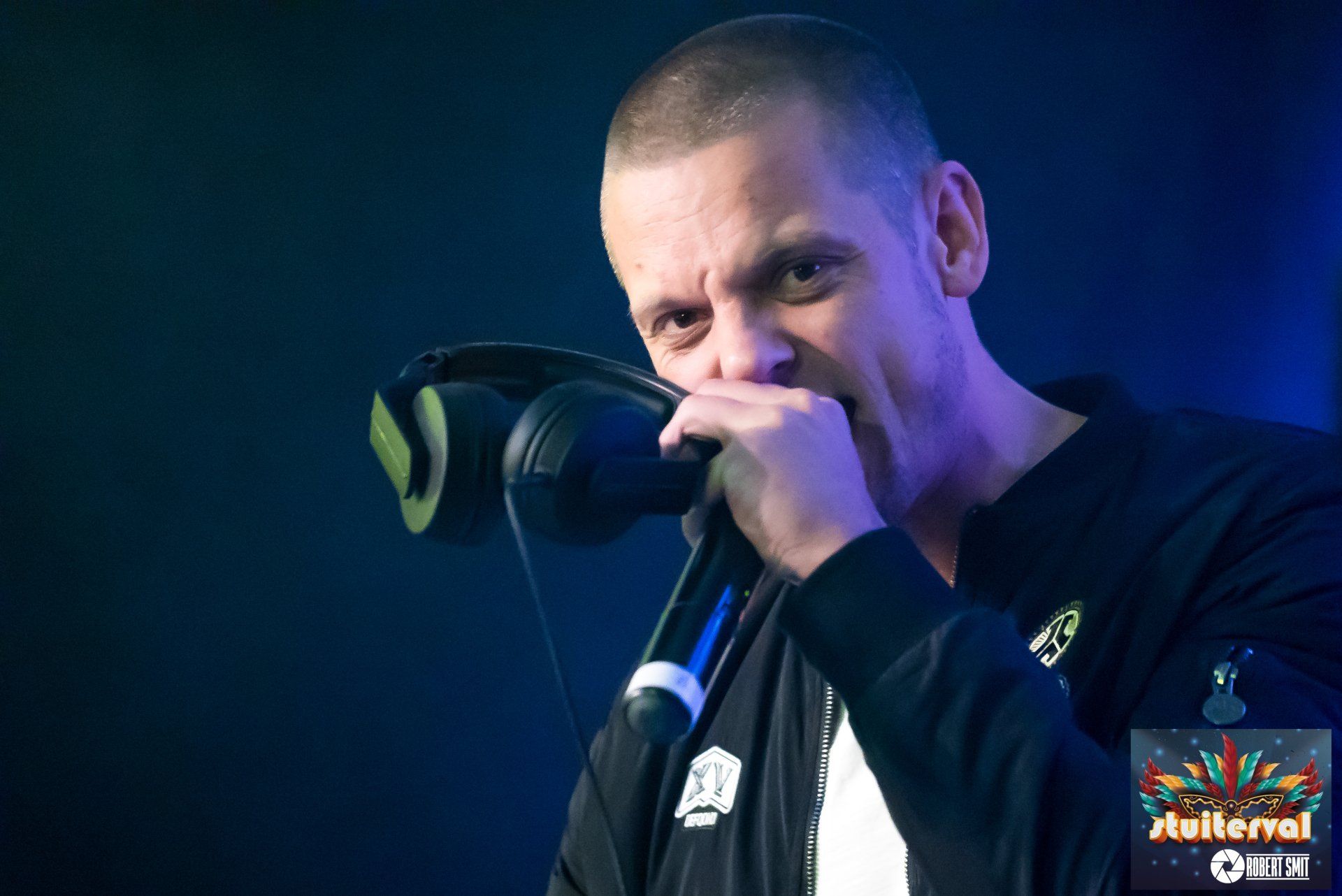 Man met geschoren hoofd rapt in een microfoon, koptelefoon om zijn nek, op een podium, donkere achtergrond.