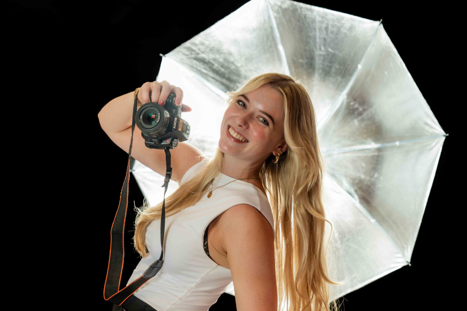 Een vrouw met lang blond haar houdt een camera vast en lacht terwijl ze voor een zilveren reflector staat tegen een zwarte achtergrond.