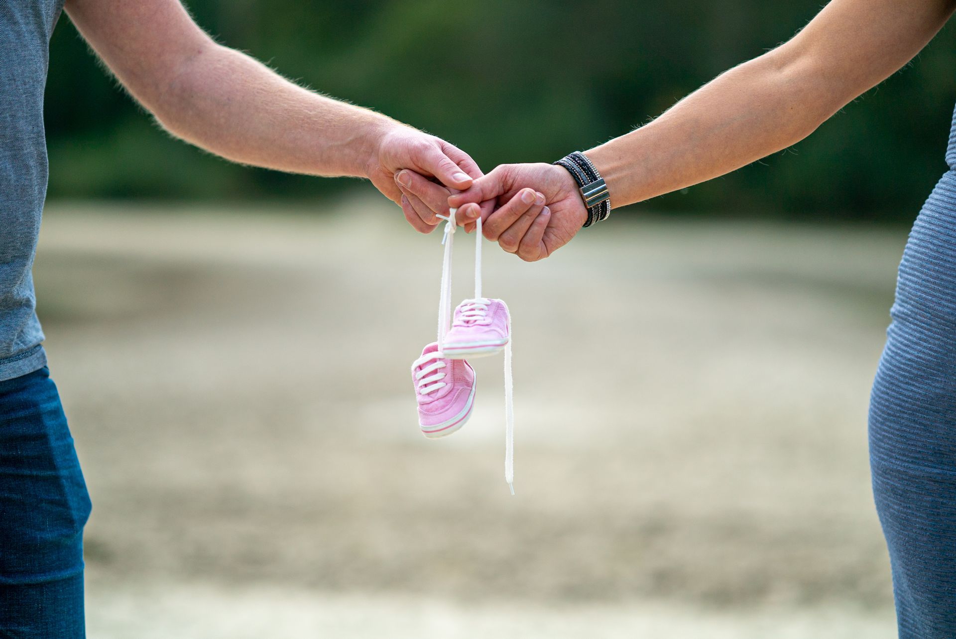 Paar dat elkaars hand vasthoudt, met roze babyschoentjes tussen hen in; wazige buitenachtergrond.