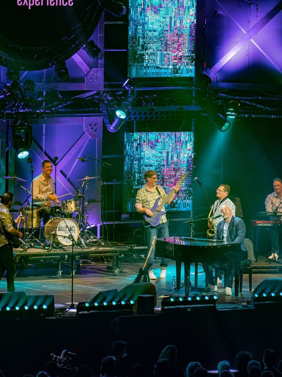 Band treedt op op een podium met kleurrijke lichten en achtergrondschermen.
