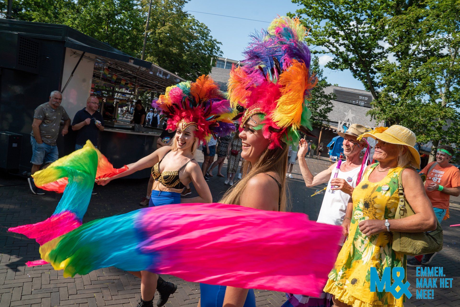Mensen op een Pride-evenement, twee van hen dragen grote, kleurrijke hoofdtooien. Een van hen houdt een regenboogsjaal vast, anderen glimlachen.