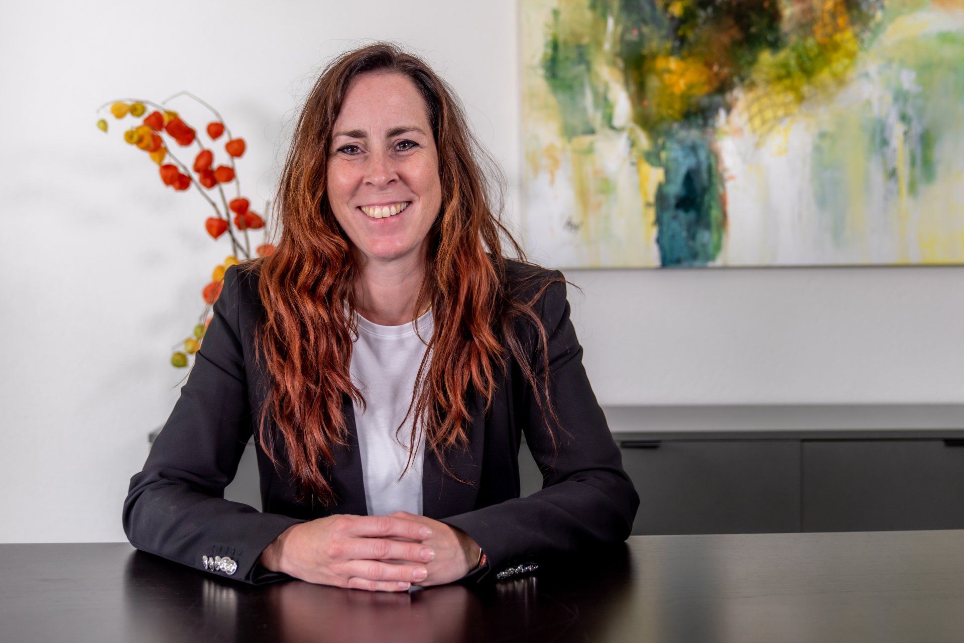 Vrouw met lang kastanjebruin haar glimlacht achter een bureau voor een abstract schilderij. Ze draagt ​​een donkere blazer en een witte blouse.