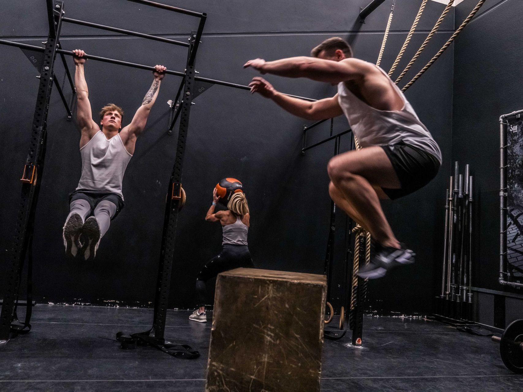 Drie mensen trainen in een donkere sportschool: een doet pull-ups, een ander springt en een derde houdt een medicijnbal vast.