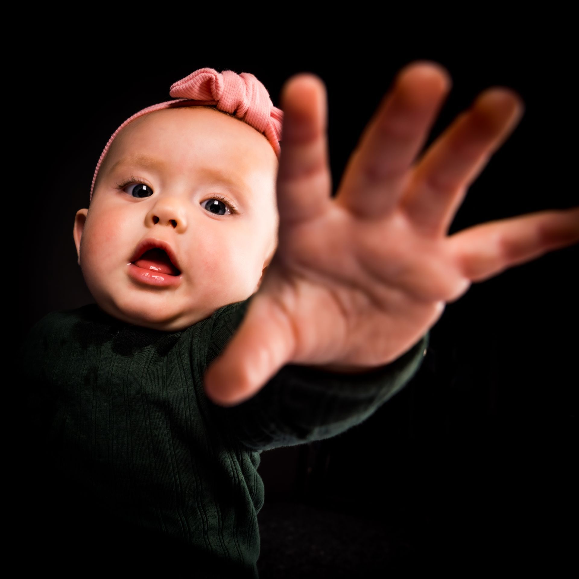 Een baby met een roze hoofdband reikt naar de kijker, met open mond. Donkergroene trui tegen een zwarte achtergrond.