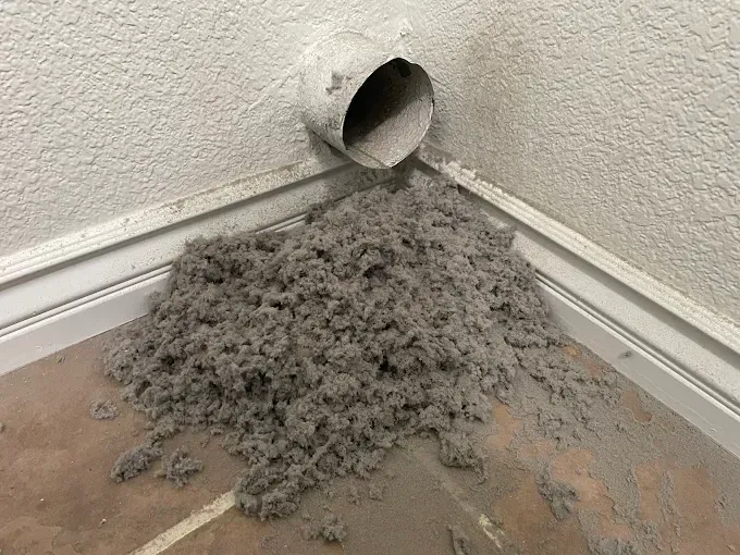 A pile of gray lint on the floor beneath a dryer vent in a corner.