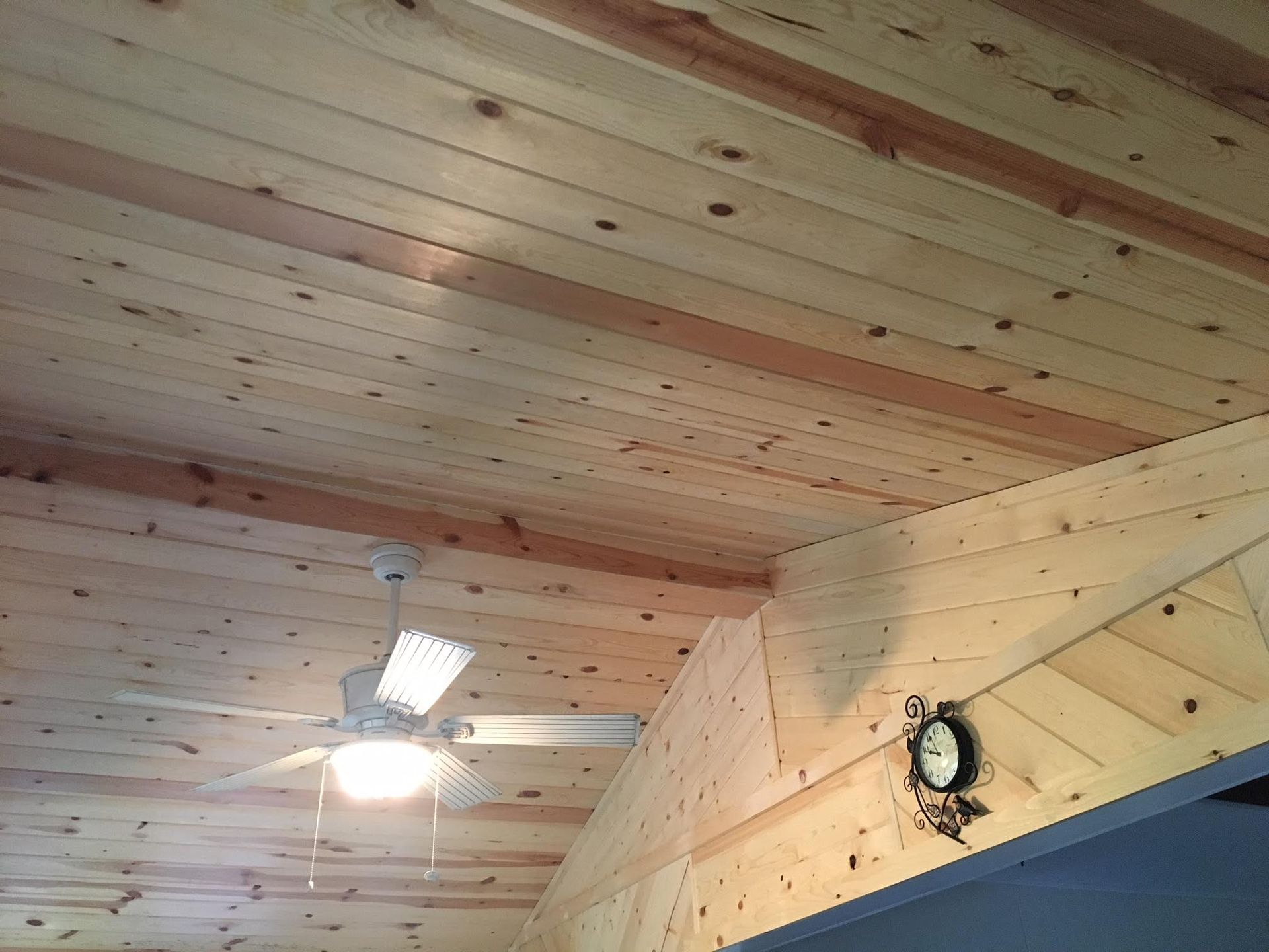 A wooden ceiling with a ceiling fan and a clock on the wall.