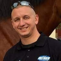 A man wearing sunglasses and a black shirt is smiling in front of a horse.