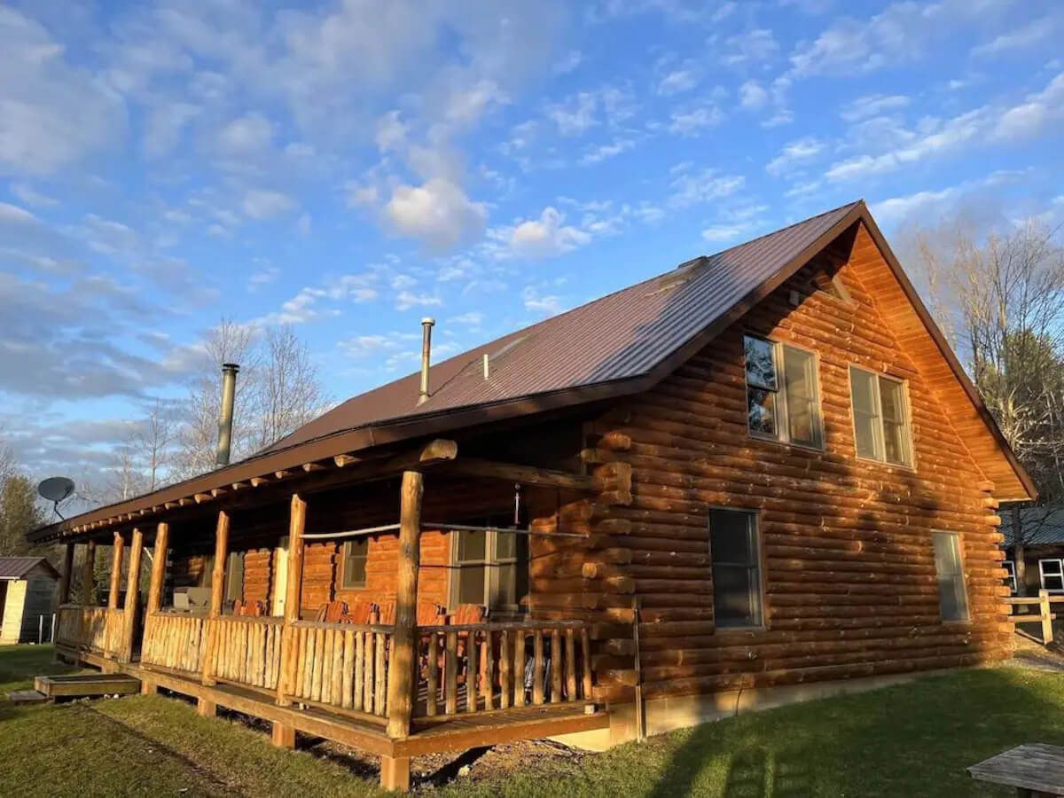 A rustic, multi-story log cabin with a wide, covered front porch under a partly cloudy blue sky.