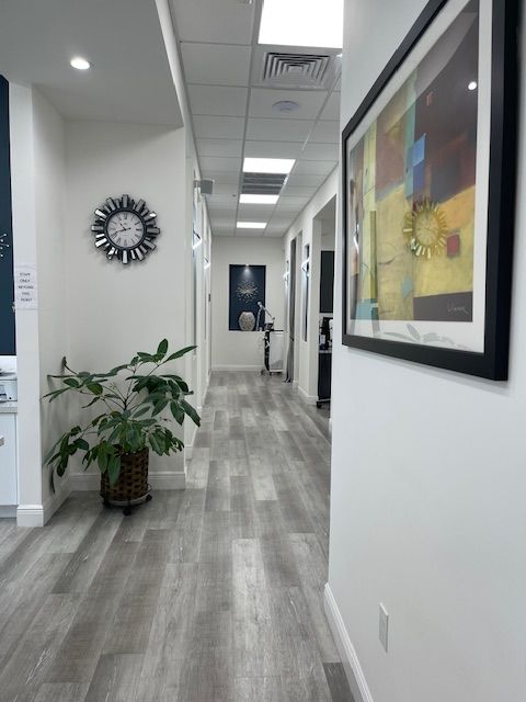 Office hallway with gray wood floors, white walls, potted plant, wall art, and a mirror at the end.