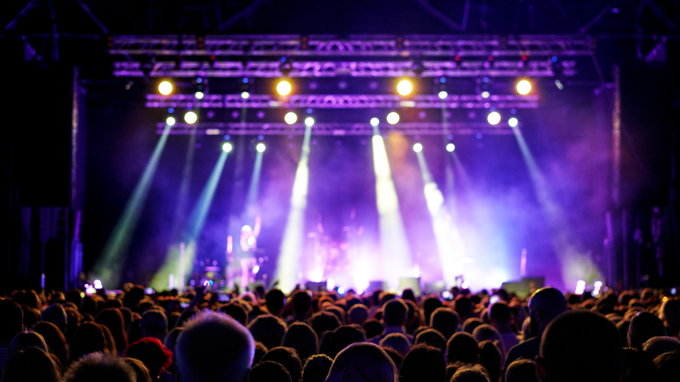 A large, silhouetted crowd watches a brightly lit concert stage with purple lighting and intense spotlights.