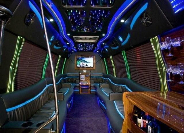 The interior of a luxury party bus with plush dark leather wrap-around seating, blue accent lighting, and a bar.