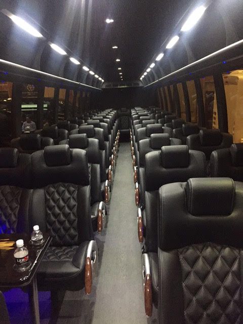 Interior view of a luxury bus with rows of black quilted leather seats facing a central aisle under recessed lighting.