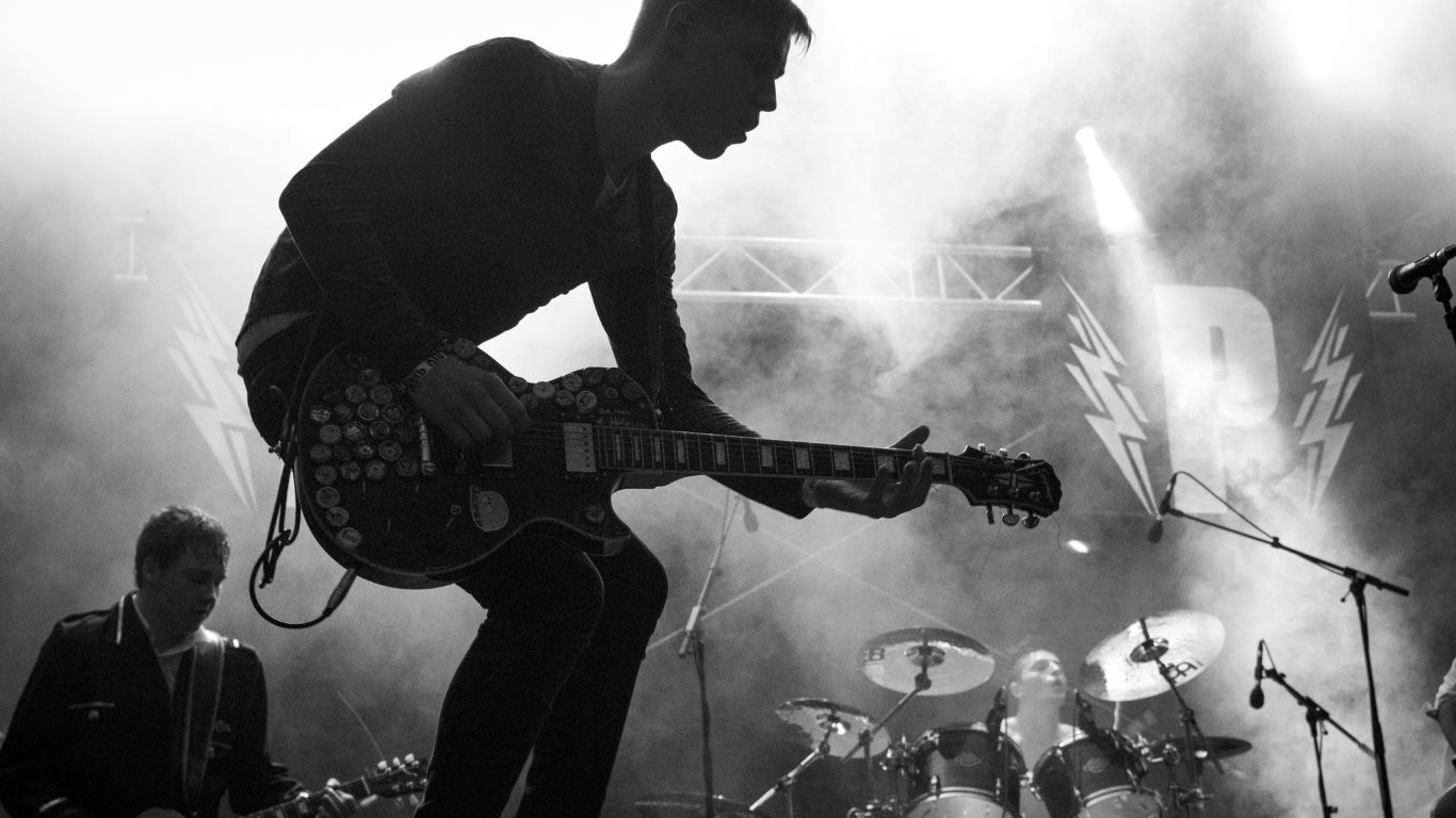 A black-and-white performance shot of a band on stage, featuring a guitarist in the foreground amid clouds of smoke.