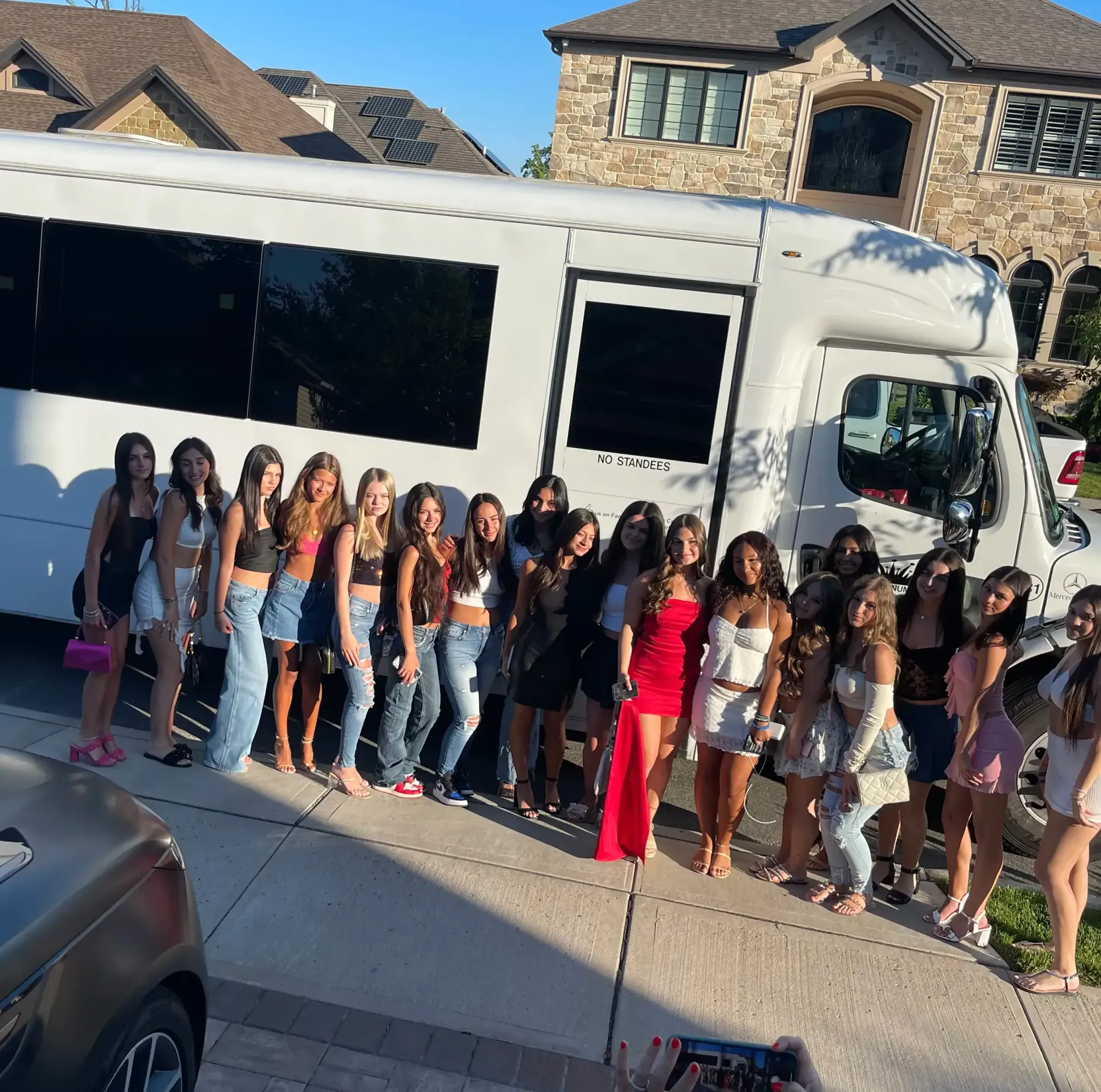 A large group posing for a photo in front of a white party bus parked on a suburban driveway.