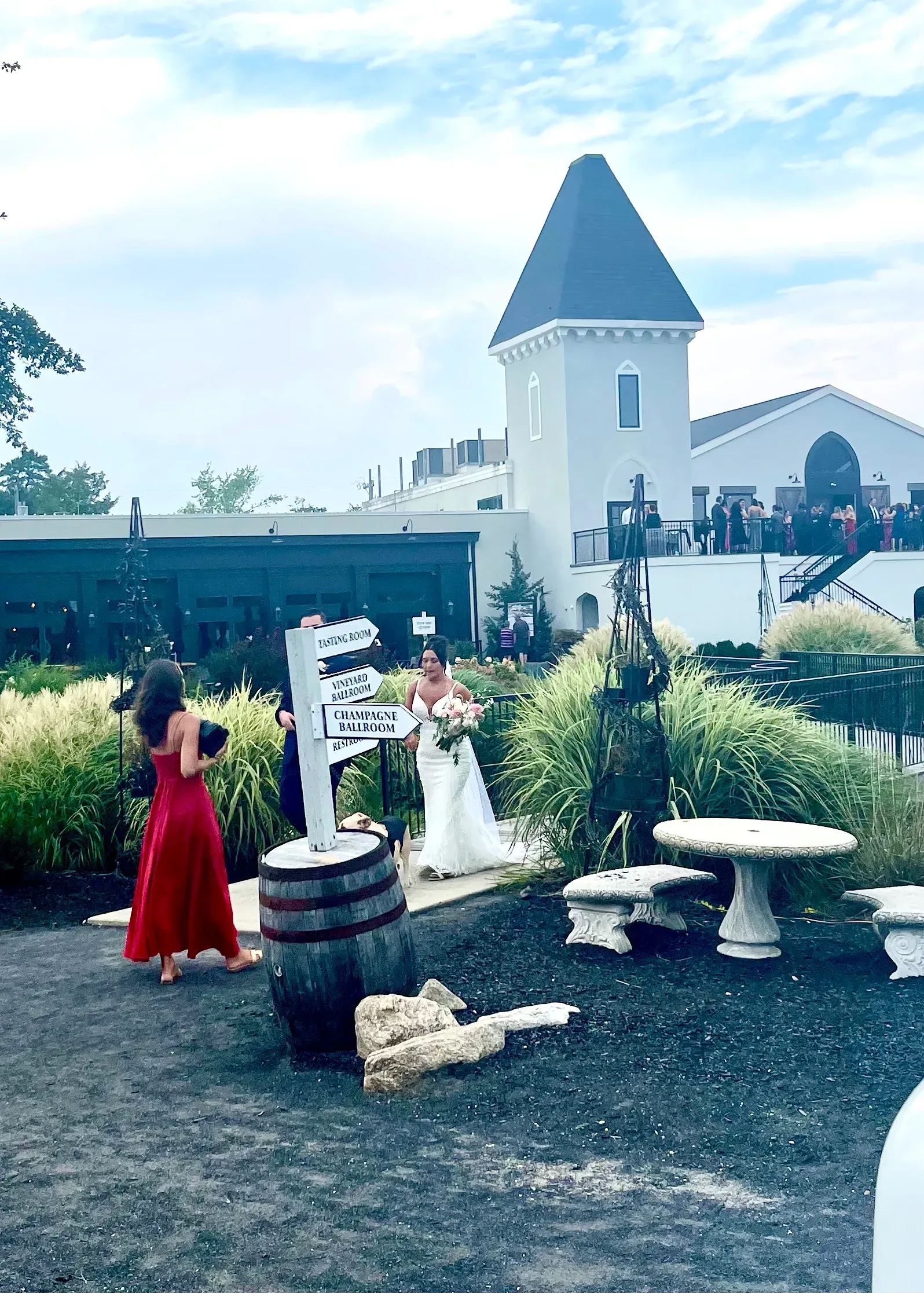 A bride in a white gown walks outdoors past a barrel sign near a castle-like building with stone furniture.