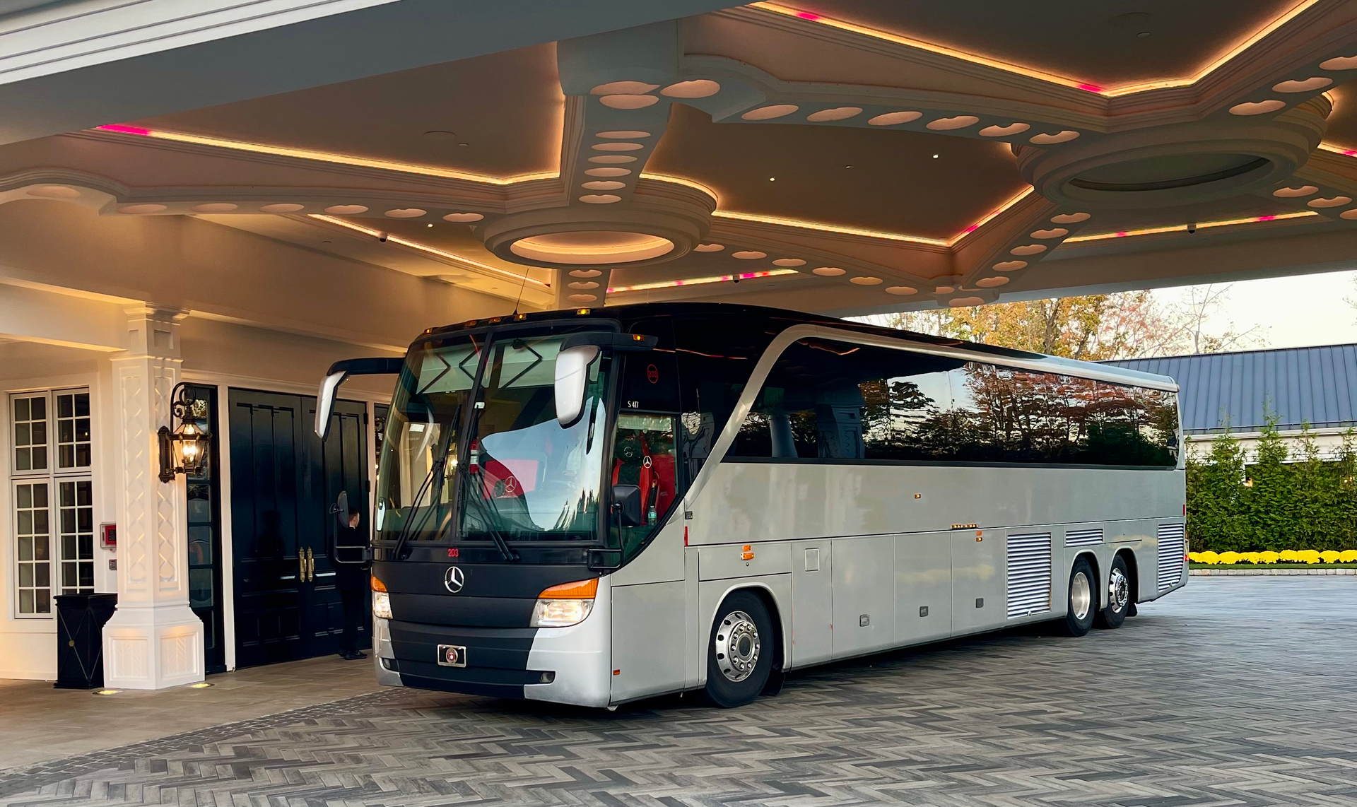 A silver tour bus is parked under the illuminated, arched canopy of a building entrance with a cobblestone driveway.