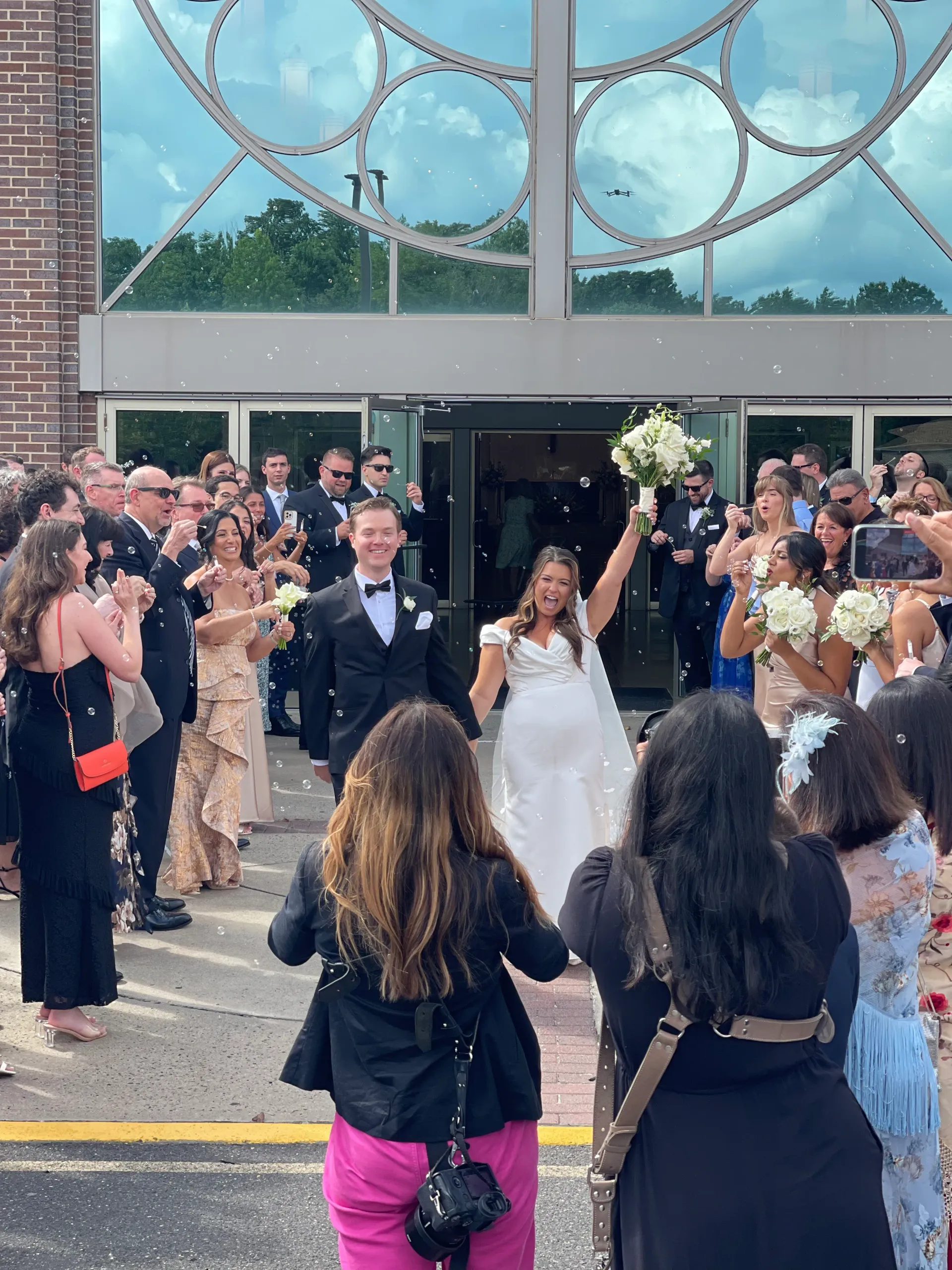A bride and groom walk hand-in-hand out of a building, surrounded by cheering guests throwing confetti at a wedding.
