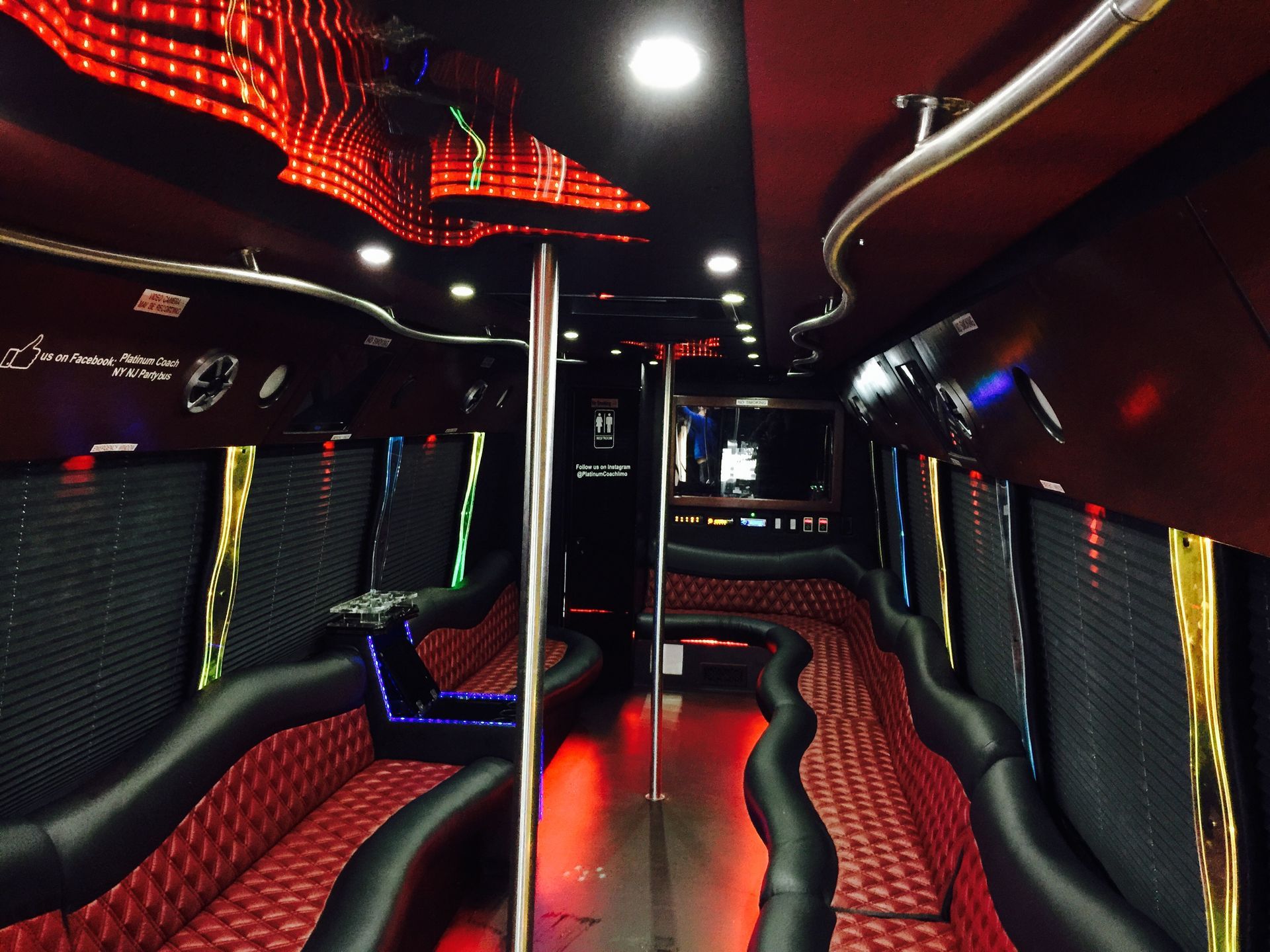 The interior of a party bus with black leather seats, red patterned upholstery, a central dance pole, and LED ceiling lights.
