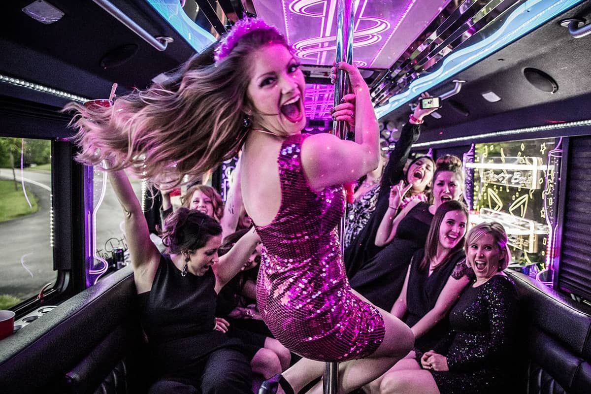 A group of people wearing party clothes celebrate in a limousine with purple lighting and a dancing pole.