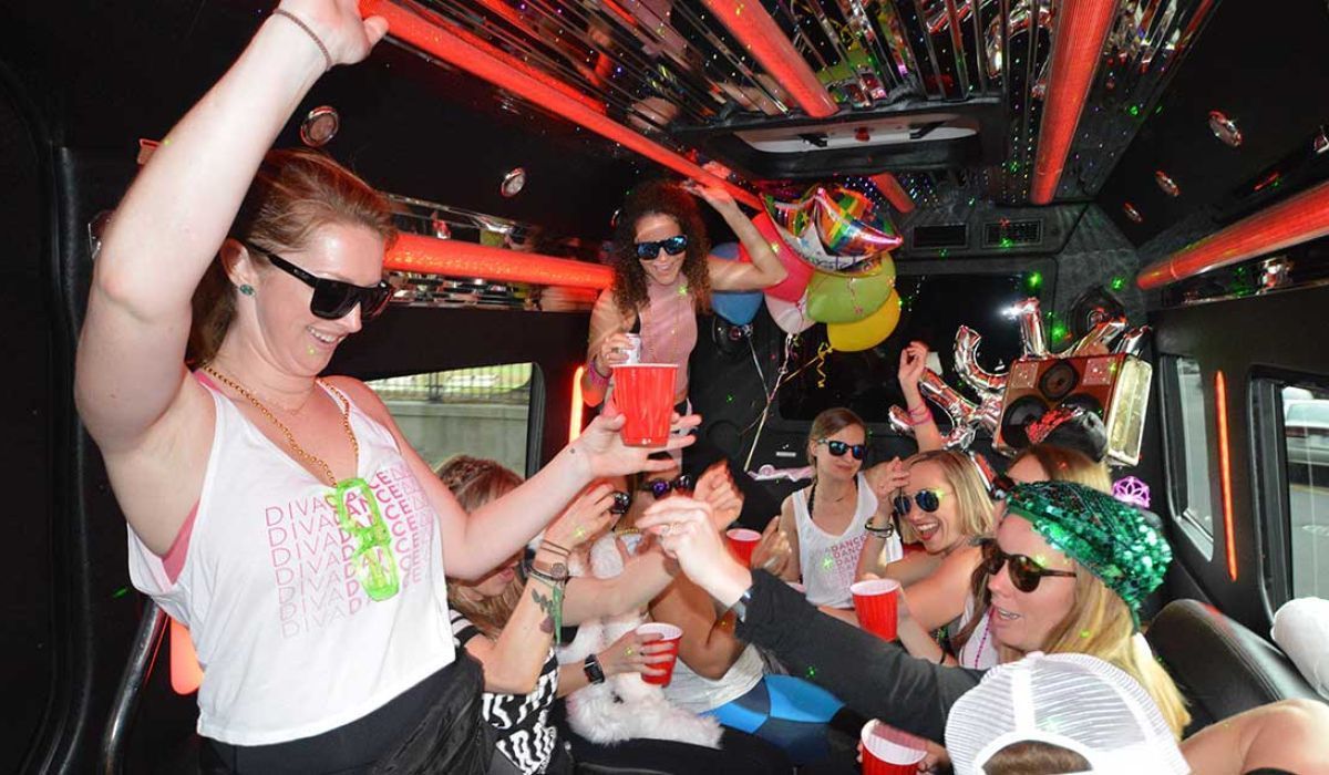A group of people wearing sunglasses and party attire enjoy drinks inside a brightly lit, neon-decorated party bus.