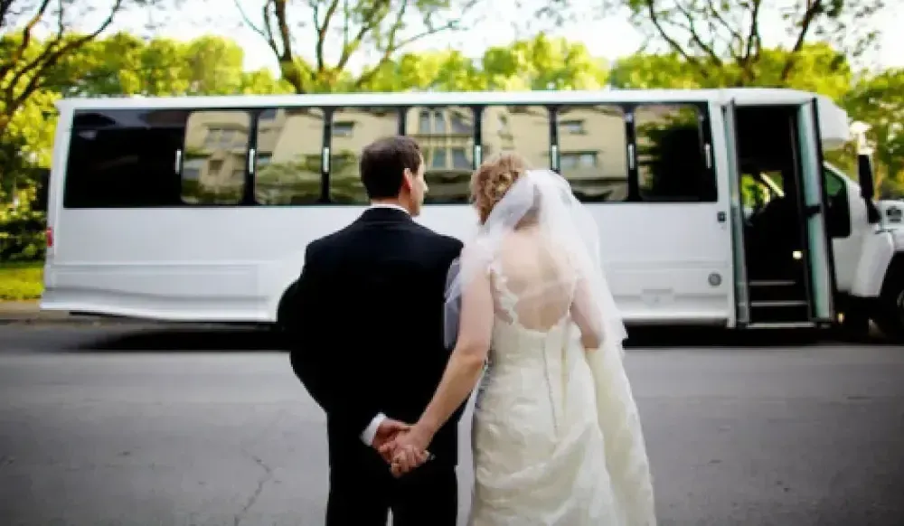 A bride and groom in wedding attire stand holding hands in front of a parked white shuttle bus on a tree-lined street.