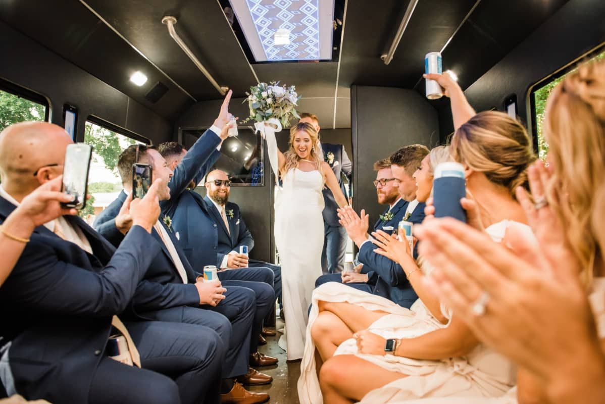 A bride holding a bouquet walks down the aisle of a party bus while guests cheer and toast with drinks.