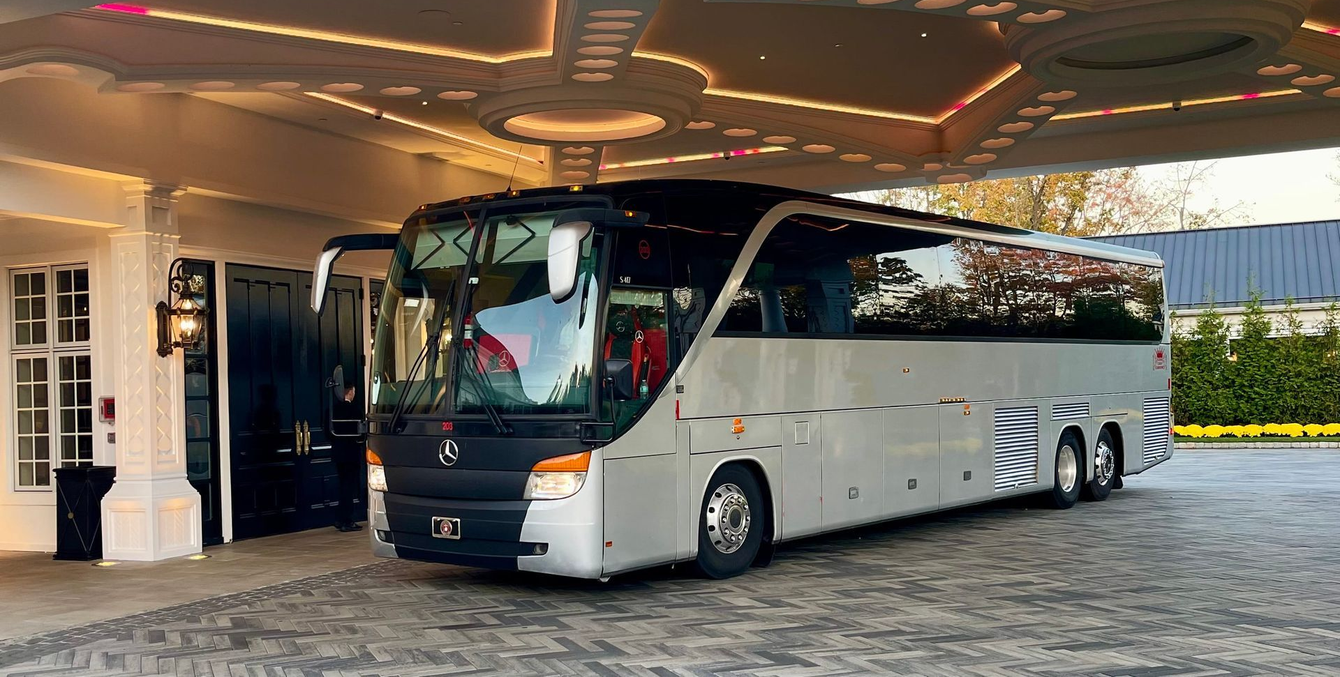 A silver Mercedes-Benz tour bus parked on a grassy field, featuring red logo decals on its side.