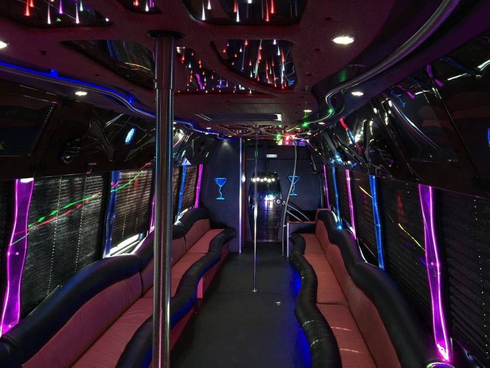 Interior of a party bus featuring pink wrap-around bench seating, neon purple lighting, and a silver dancing pole.