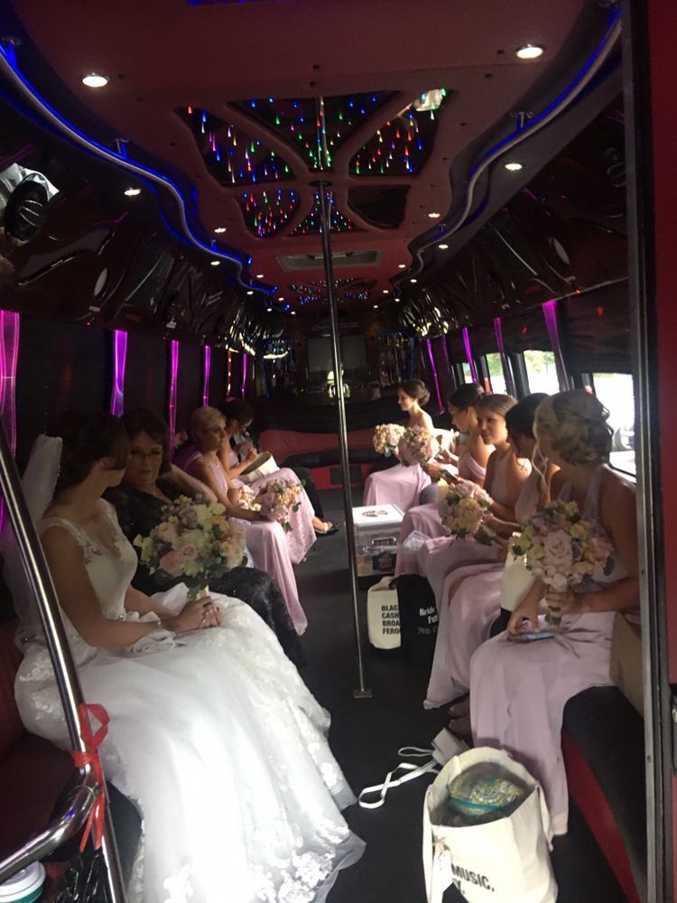 A bride and bridesmaids in formal attire holding bouquets inside a dimly lit limousine with purple accent lighting.