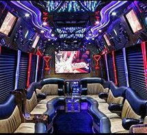 Luxury limousine interior with plush tan and black curved leather seating, blue ambient lighting, and a large TV screen.