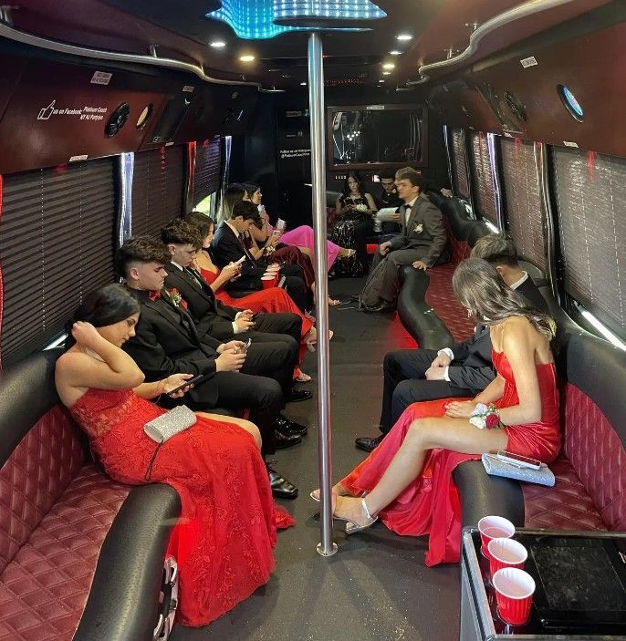 A group of people in formal attire sits inside a dark, lit party bus, many looking down at their phones.