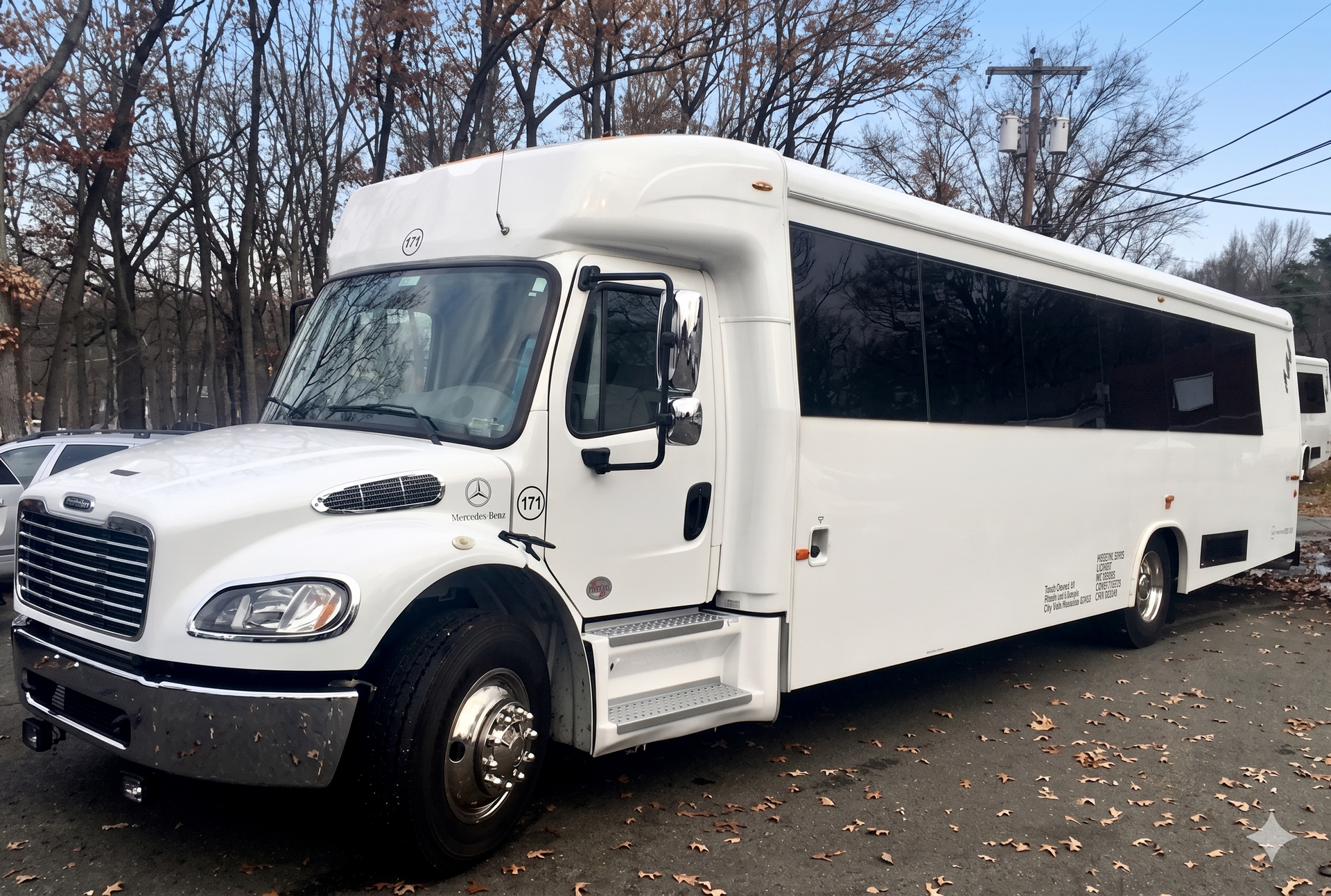 A white Freightliner passenger shuttle bus parked on a paved lot with a wooded background.